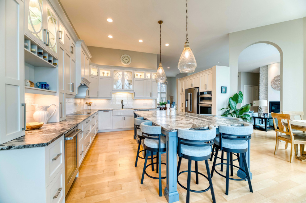 Large transitional l-shaped light wood floor and brown floor eat-in kitchen photo with a farmhouse sink, shaker cabinets, white cabinets, solid surface countertops, ceramic backsplash, stainless steel appliances and an island