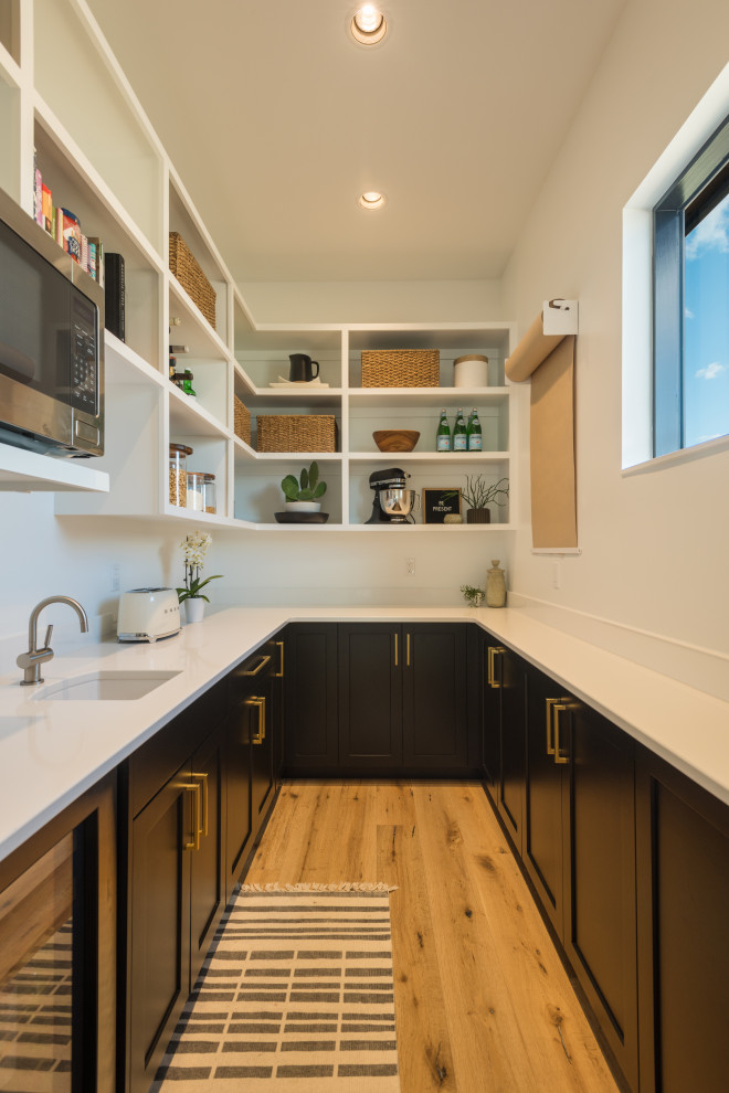 Cette photo montre une cuisine chic avec un évier de ferme, un placard à porte shaker, des portes de placard noires, un plan de travail en quartz modifié, un électroménager en acier inoxydable, un sol en bois brun, îlot et un plan de travail blanc.