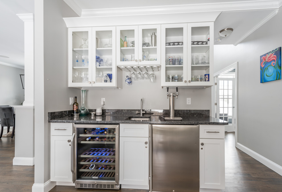 This is an example of a mid-sized transitional single-wall wet bar in New York with an undermount sink, glass-front cabinets, white cabinets, granite benchtops, dark hardwood floors and brown floor.
