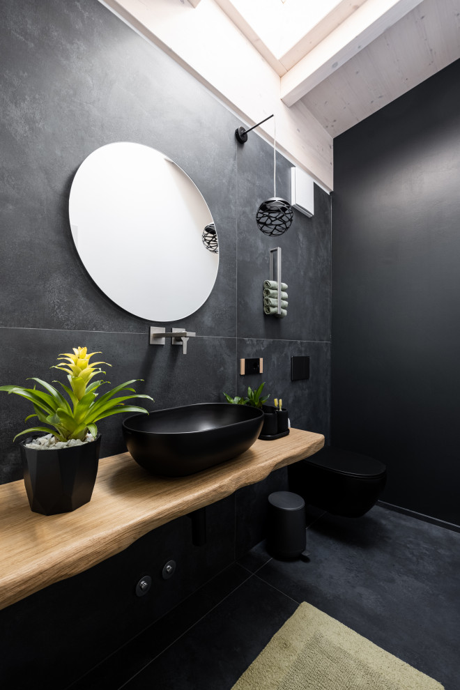 На фото: туалет в современном стиле с светлыми деревянными фасадами, черной плиткой, черными стенами, полом из керамической плитки, настольной раковиной, столешницей из дерева, черным полом, подвесной тумбой и балками на потолке
