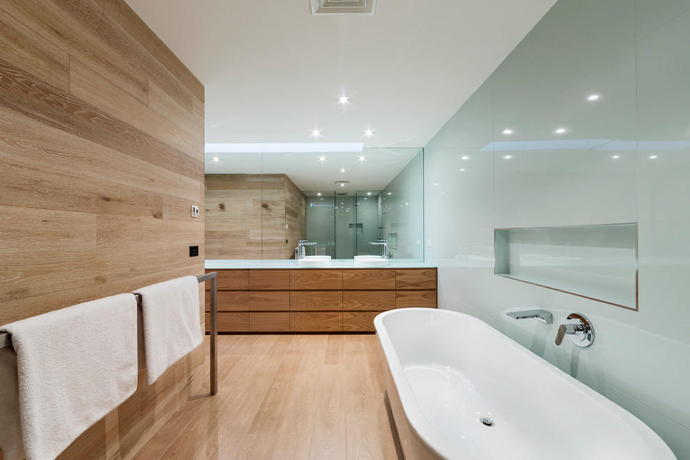 This is an example of a modern bathroom in Melbourne with a vessel sink, light wood cabinets, glass benchtops, a freestanding tub, green tile, glass tile and light hardwood floors.