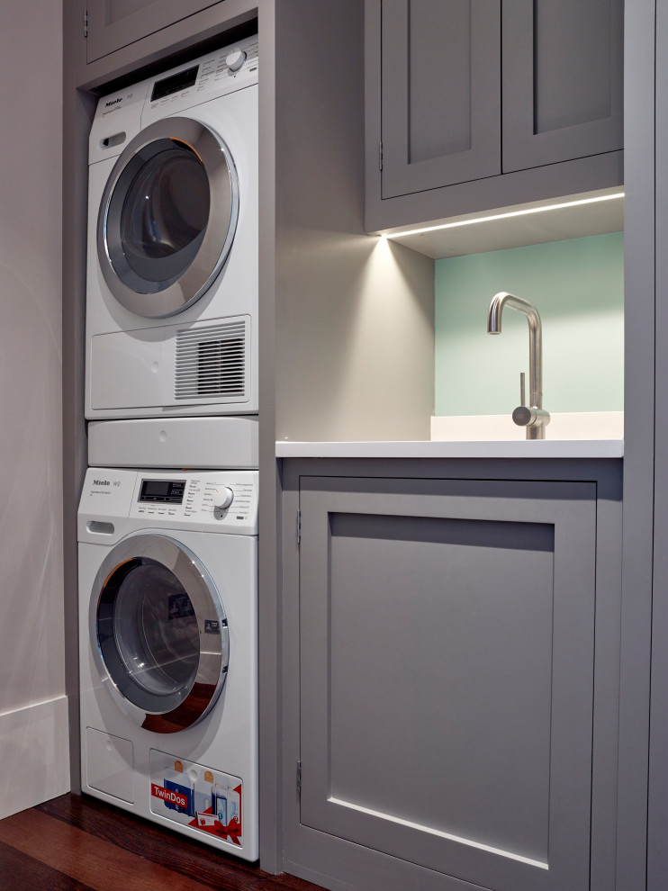 Small contemporary single-wall dedicated laundry room in London with an integrated sink, shaker cabinets, grey cabinets, quartzite benchtops, white walls, dark hardwood floors, a stacked washer and dryer, brown floor and white benchtop.