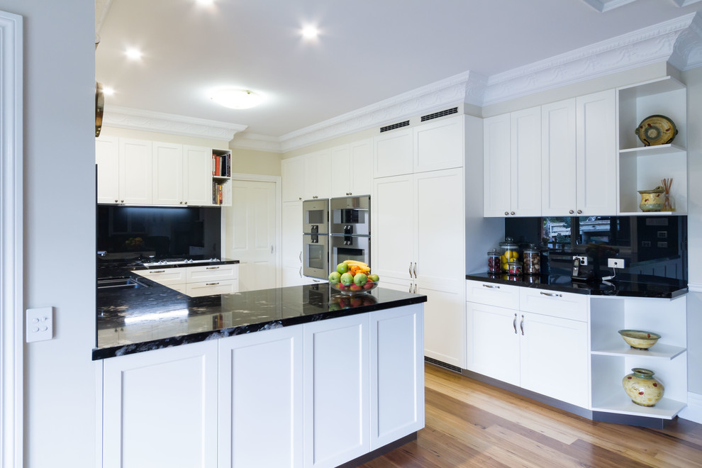 Photo of a large traditional u-shaped open plan kitchen in Melbourne with an undermount sink, shaker cabinets, white cabinets, granite benchtops, black splashback, ceramic splashback, stainless steel appliances, light hardwood floors and brown floor.