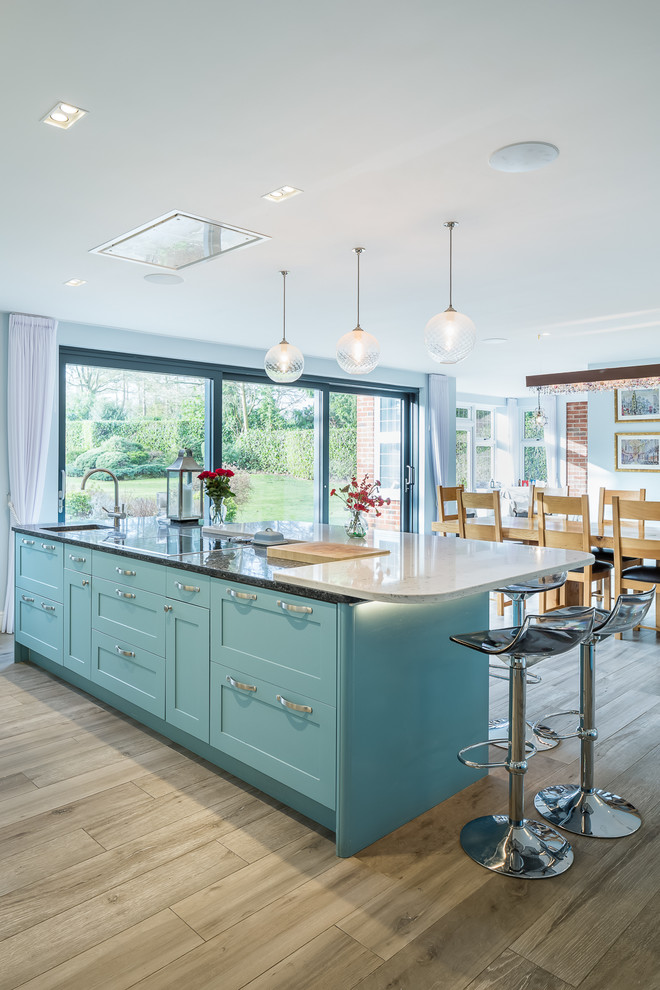 This is an example of a large contemporary galley open plan kitchen in Hertfordshire with a drop-in sink, shaker cabinets, blue cabinets, granite benchtops, white splashback, stone slab splashback, stainless steel appliances, medium hardwood floors, multiple islands, brown floor and black benchtop.
