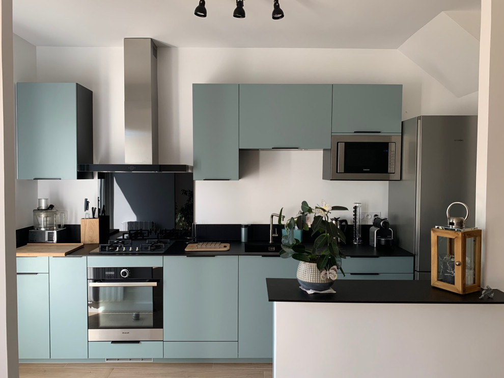 Photo of a mid-sized modern galley open plan kitchen with an undermount sink, flat-panel cabinets, green cabinets, granite benchtops, black splashback, granite splashback, stainless steel appliances, light hardwood floors, with island, brown floor, black benchtop and wallpaper.