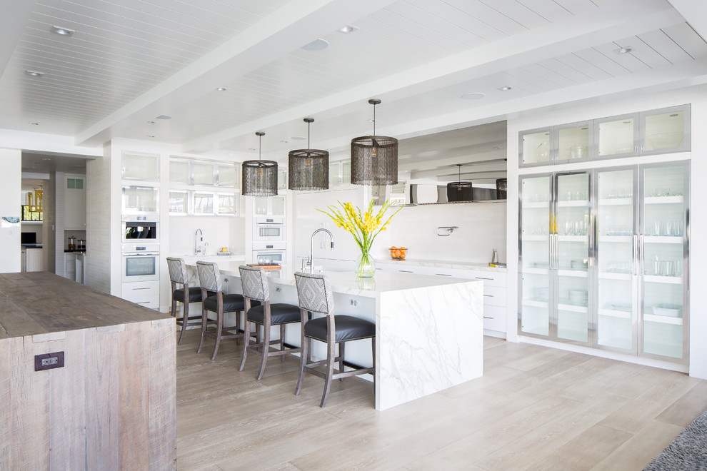 Design ideas for a beach style l-shaped kitchen in Orange County with an undermount sink, flat-panel cabinets, white cabinets, white appliances, light hardwood floors, with island, beige floor and white benchtop.