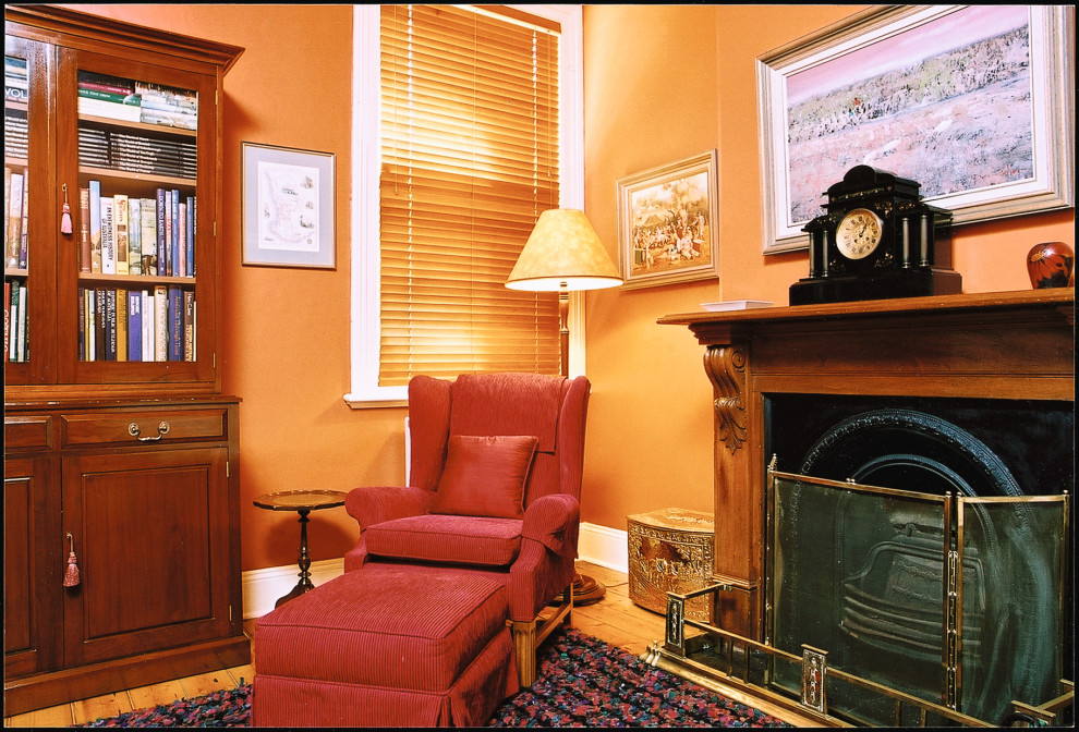 Klassisk inredning av ett litet arbetsrum, med ett bibliotek, röda väggar, ljust trägolv, en standard öppen spis, en spiselkrans i trä, ett fristående skrivbord och brunt golv