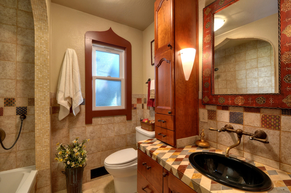 This is an example of a small mediterranean bathroom in San Francisco with a drop-in sink, medium wood cabinets, tile benchtops, an alcove tub, a shower/bathtub combo, a one-piece toilet, multi-coloured tile, ceramic tile, beige walls, travertine floors and flat-panel cabinets.