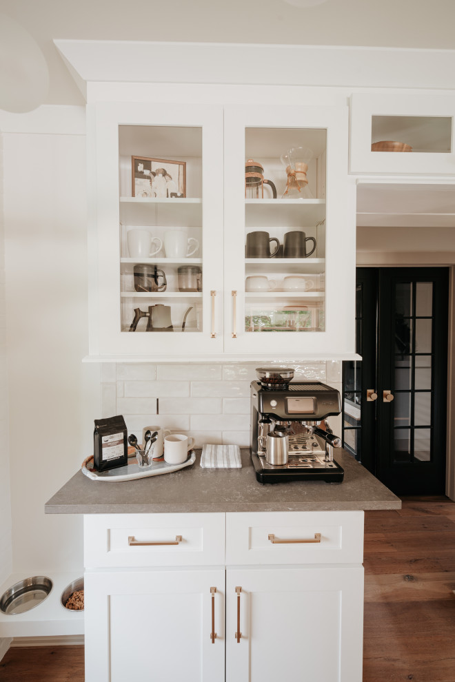 This is an example of a modern galley eat-in kitchen in Detroit with an undermount sink, glass-front cabinets, white cabinets, quartz benchtops, white splashback, porcelain splashback, white appliances, light hardwood floors, with island, brown floor and multi-coloured benchtop.