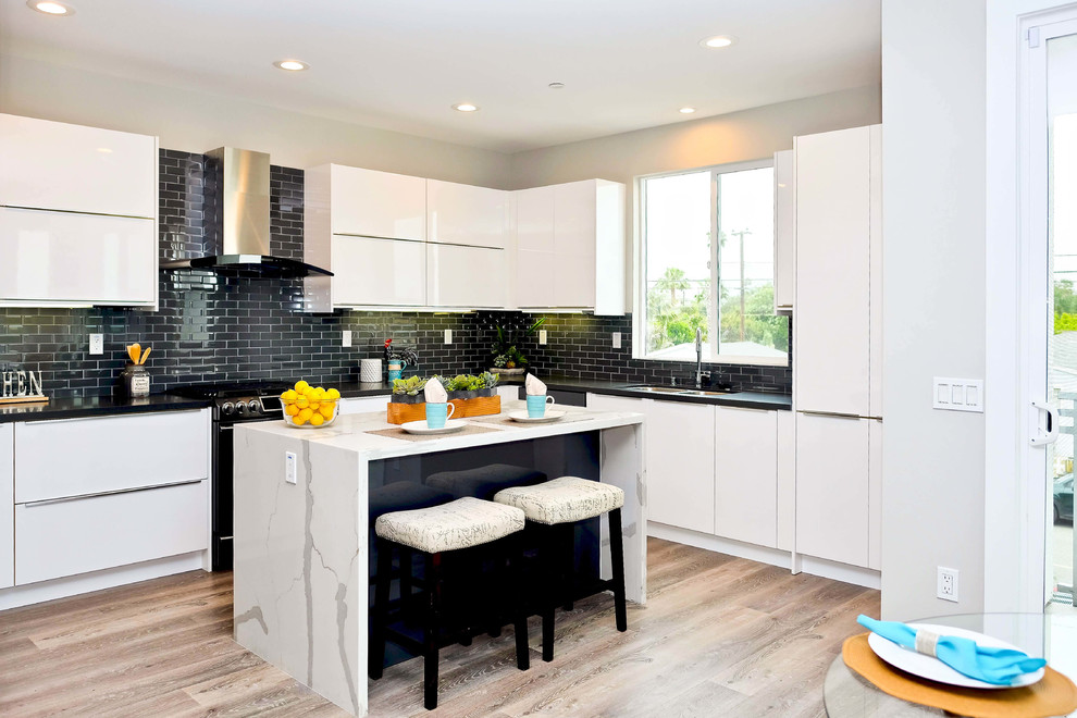 This is an example of a mid-sized contemporary u-shaped eat-in kitchen in Orange County with flat-panel cabinets, white cabinets, quartz benchtops, black splashback, ceramic splashback, black appliances, light hardwood floors, with island, brown floor, black benchtop and a double-bowl sink.