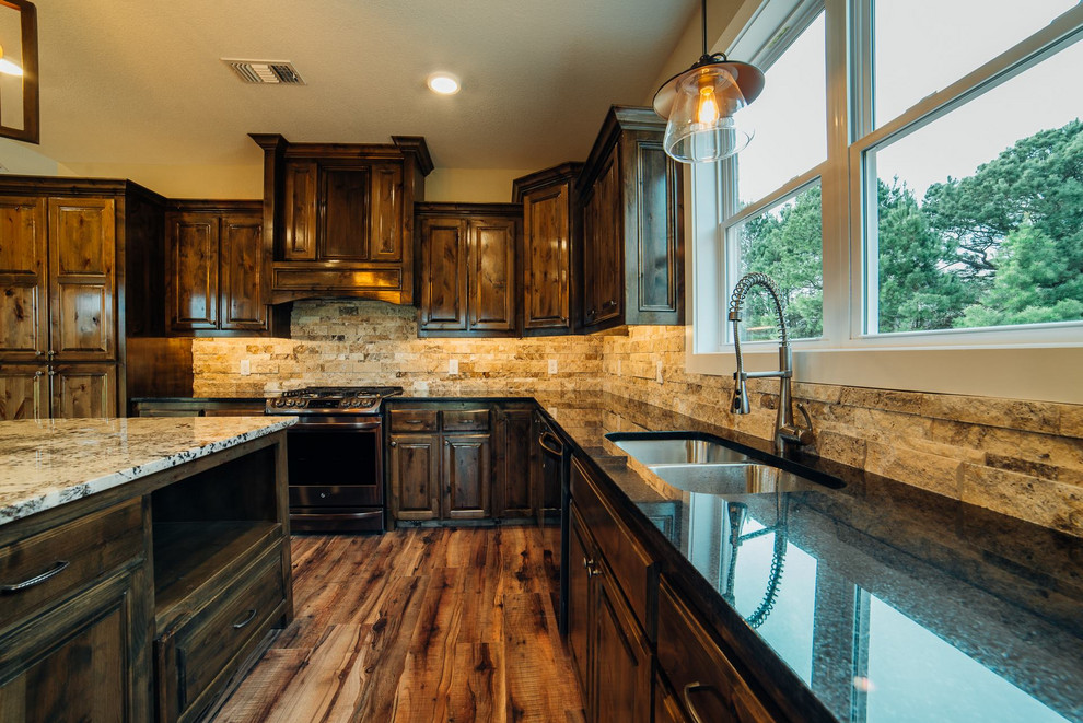 Inspiration for a large country l-shaped open plan kitchen in Dallas with a double-bowl sink, raised-panel cabinets, dark wood cabinets, granite benchtops, beige splashback, travertine splashback, stainless steel appliances, vinyl floors, with island, beige floor and black benchtop.