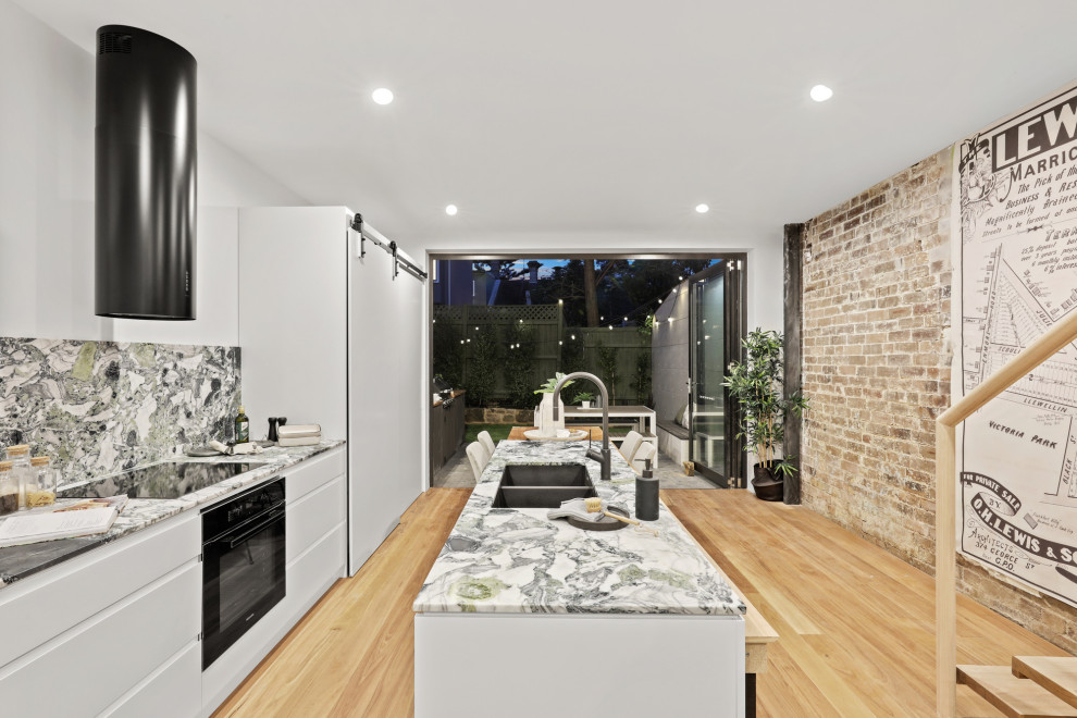Photo of a contemporary galley kitchen in Sydney with an undermount sink, flat-panel cabinets, white cabinets, multi-coloured splashback, stone slab splashback, black appliances, light hardwood floors, with island, beige floor, multi-coloured benchtop and marble benchtops.