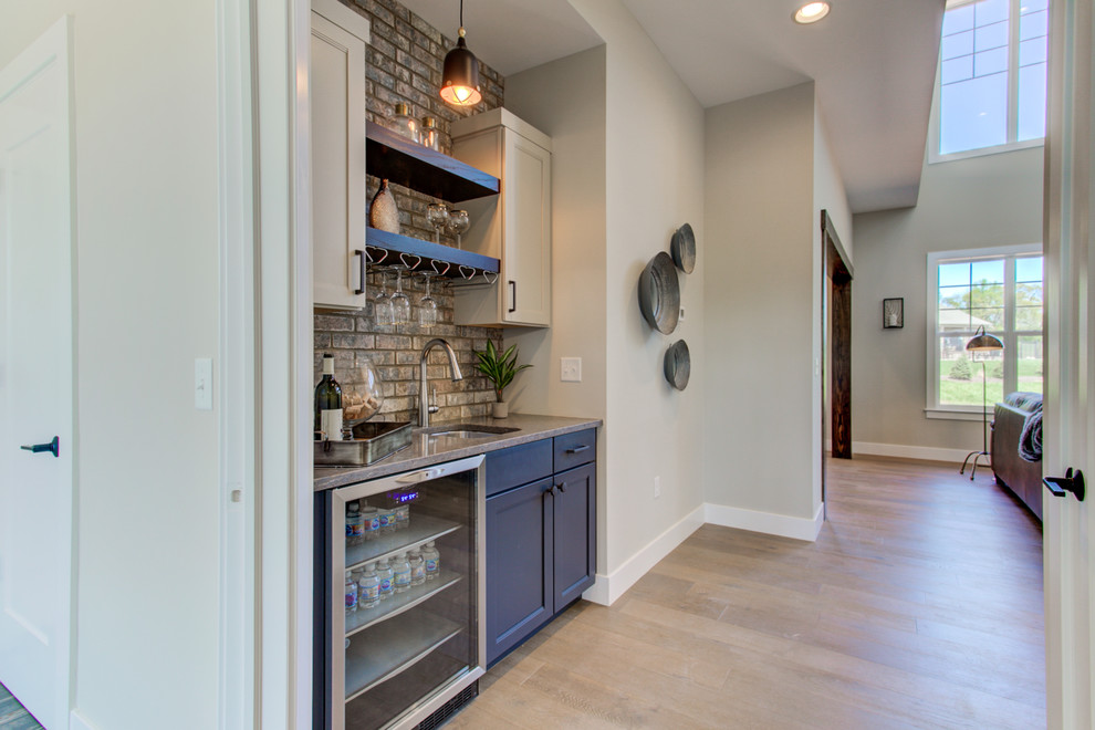 This is an example of a large arts and crafts single-wall wet bar in Other with an undermount sink, blue cabinets, quartz benchtops, beige splashback, brick splashback, medium hardwood floors and beige benchtop.