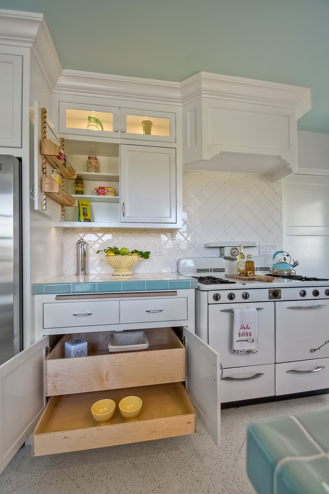 This is an example of a mid-sized transitional galley separate kitchen in San Diego with a double-bowl sink, recessed-panel cabinets, white cabinets, tile benchtops, white splashback, ceramic splashback, white appliances, terrazzo floors and no island.