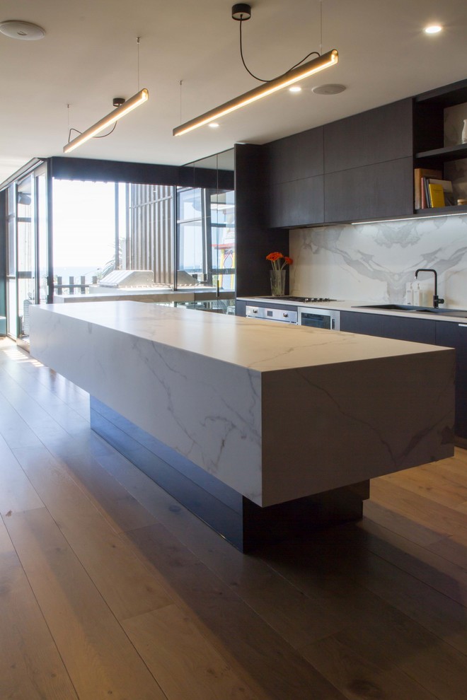 This is an example of a mid-sized modern galley open plan kitchen in Melbourne with flat-panel cabinets, with island, a drop-in sink, black cabinets, tile benchtops, white splashback, porcelain splashback, stainless steel appliances, medium hardwood floors, brown floor and white benchtop.