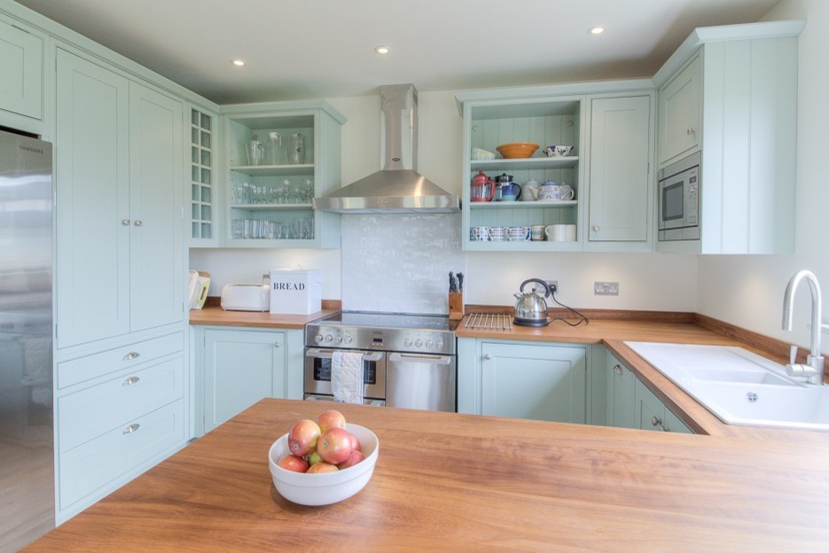 This is an example of a mid-sized beach style u-shaped open plan kitchen in London with a double-bowl sink, shaker cabinets, green cabinets, wood benchtops, white splashback, subway tile splashback, stainless steel appliances, light hardwood floors and no island.