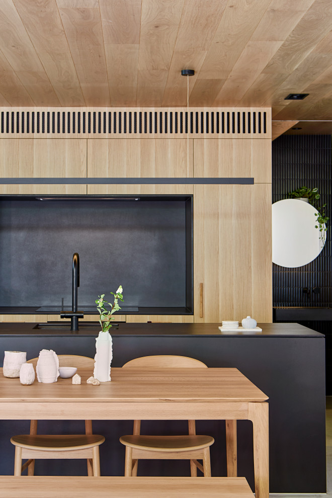Photo of a mid-sized industrial galley eat-in kitchen in Melbourne with an undermount sink, flat-panel cabinets, light wood cabinets, quartz benchtops, black splashback, engineered quartz splashback, black appliances, light hardwood floors, with island and black benchtop.