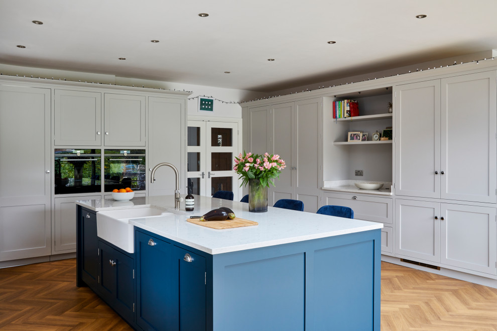 This is an example of an expansive transitional u-shaped open plan kitchen in Kent with with island, white benchtop, a farmhouse sink, shaker cabinets, grey cabinets, panelled appliances, medium hardwood floors and beige floor.