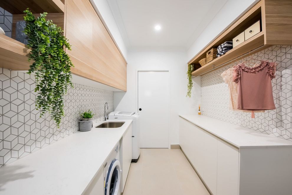 Inspiration for a mid-sized contemporary galley dedicated laundry room in Other with an undermount sink, flat-panel cabinets, white walls, a side-by-side washer and dryer, white benchtop, light wood cabinets, granite benchtops, white splashback, ceramic splashback, ceramic floors and white floor.