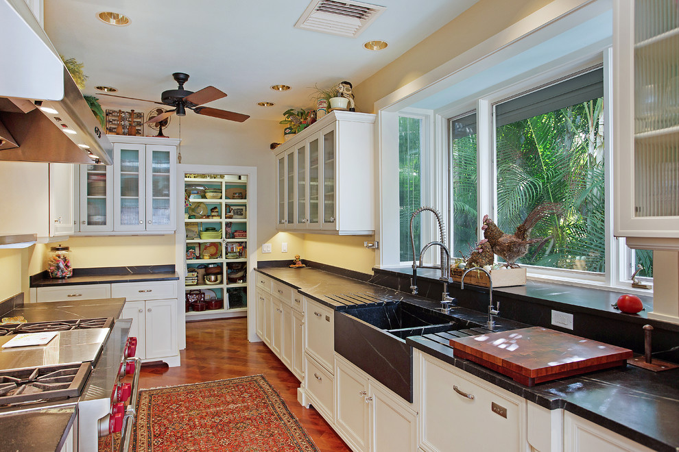 This is an example of a mid-sized traditional galley eat-in kitchen in Tampa with a farmhouse sink, recessed-panel cabinets, white cabinets, soapstone benchtops, beige splashback, stone slab splashback, stainless steel appliances, medium hardwood floors and with island.