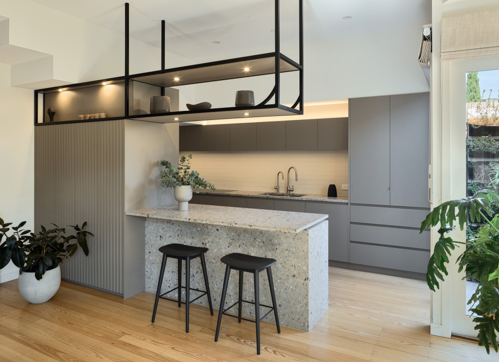 Small contemporary eat-in kitchen in Melbourne with a double-bowl sink, flat-panel cabinets, grey cabinets, terrazzo benchtops, grey splashback, ceramic splashback, black appliances, light hardwood floors, with island and multi-coloured benchtop.