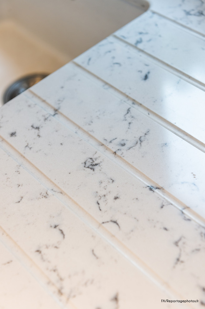 Cette photo montre une cuisine ouverte linéaire tendance de taille moyenne avec un évier intégré, un placard à porte affleurante, des portes de placard grises, un plan de travail en quartz, une crédence grise, une crédence en carrelage de pierre, un électroménager noir, un sol en carrelage de céramique, îlot, un sol beige et un plan de travail gris.