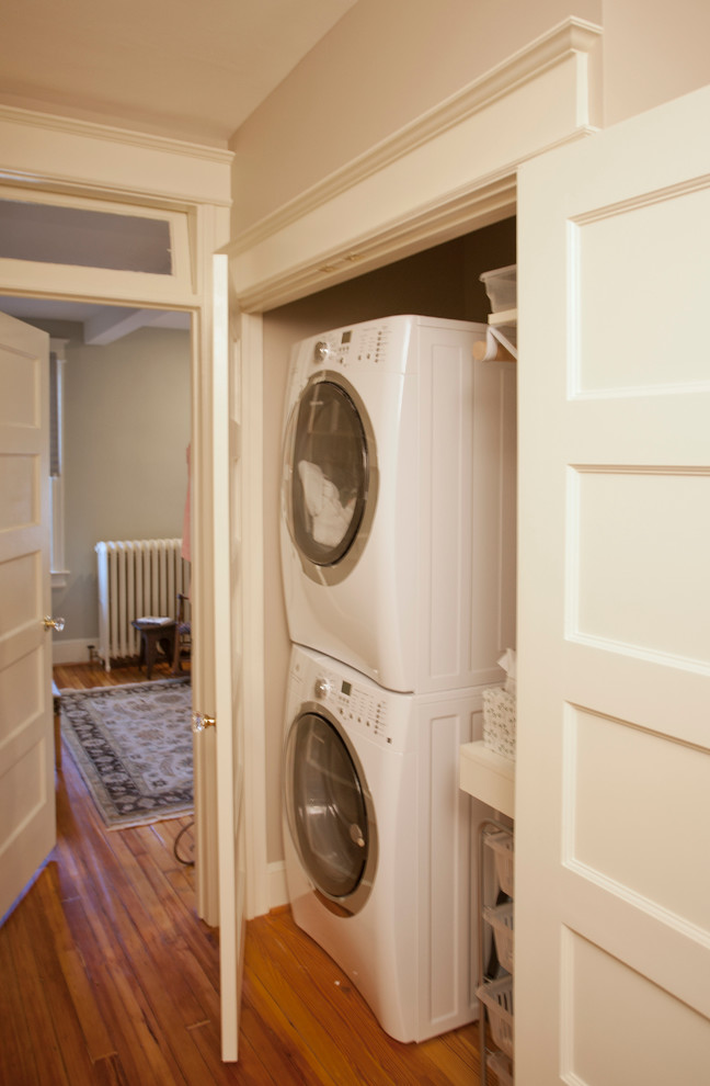 Photo of a small transitional laundry cupboard in DC Metro with white cabinets, beige walls, medium hardwood floors and a stacked washer and dryer.