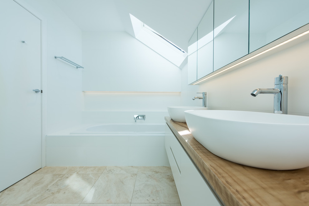 Inspiration for a mid-sized contemporary master bathroom in Sydney with flat-panel cabinets, white cabinets, a drop-in tub, an open shower, a wall-mount toilet, white tile, ceramic tile, white walls, limestone floors, a vessel sink, wood benchtops and beige floor.