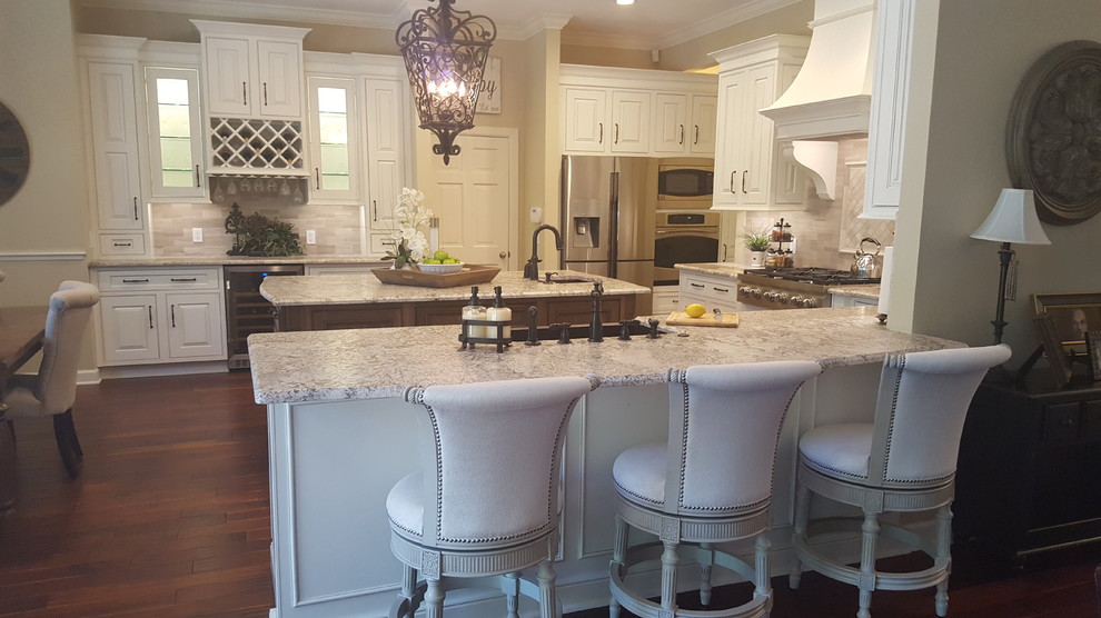 This is an example of a large transitional u-shaped open plan kitchen in Orlando with a farmhouse sink, white cabinets, granite benchtops, beige splashback, stone tile splashback, stainless steel appliances, medium hardwood floors, with island, brown floor and raised-panel cabinets.