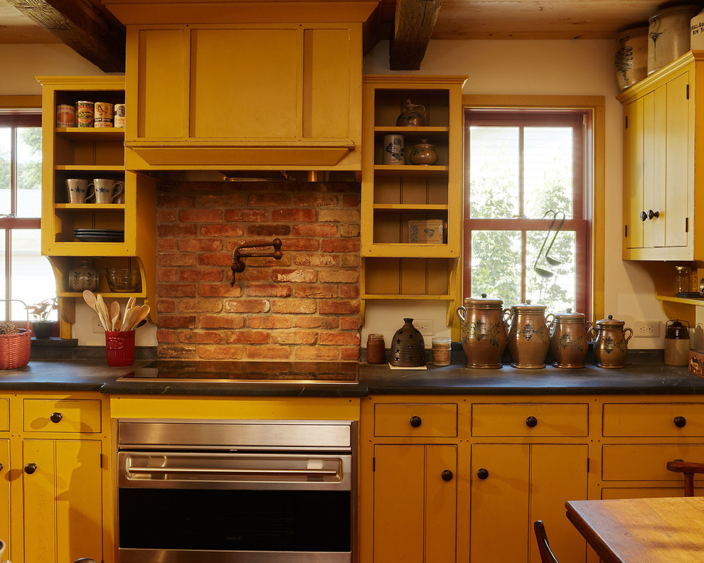 This is an example of a mid-sized country separate kitchen in New York with an integrated sink, flat-panel cabinets, yellow cabinets, soapstone benchtops, red splashback, brick splashback, stainless steel appliances, medium hardwood floors, no island, brown floor, black benchtop and exposed beam.