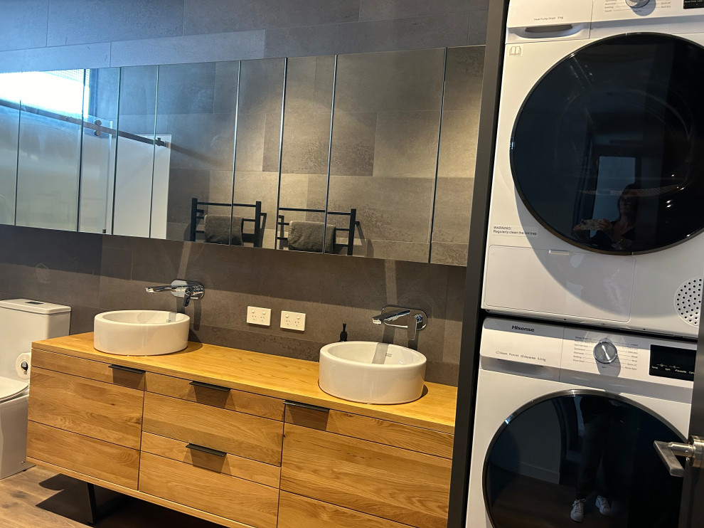 Photo of an urban bathroom in Hamilton with medium wood cabinets, a double shower, a one-piece toilet, grey tiles, mirror tiles, multi-coloured walls, cork flooring, a trough sink, wooden worktops, multi-coloured floors, a sliding door, a laundry area, double sinks and a freestanding vanity unit.