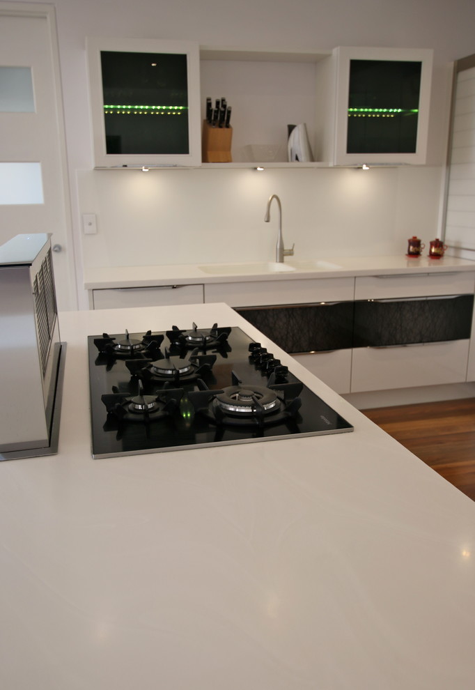 Design ideas for a large modern l-shaped open plan kitchen in Perth with an integrated sink, flat-panel cabinets, white cabinets, solid surface benchtops, white splashback, glass sheet splashback, black appliances, medium hardwood floors and with island.
