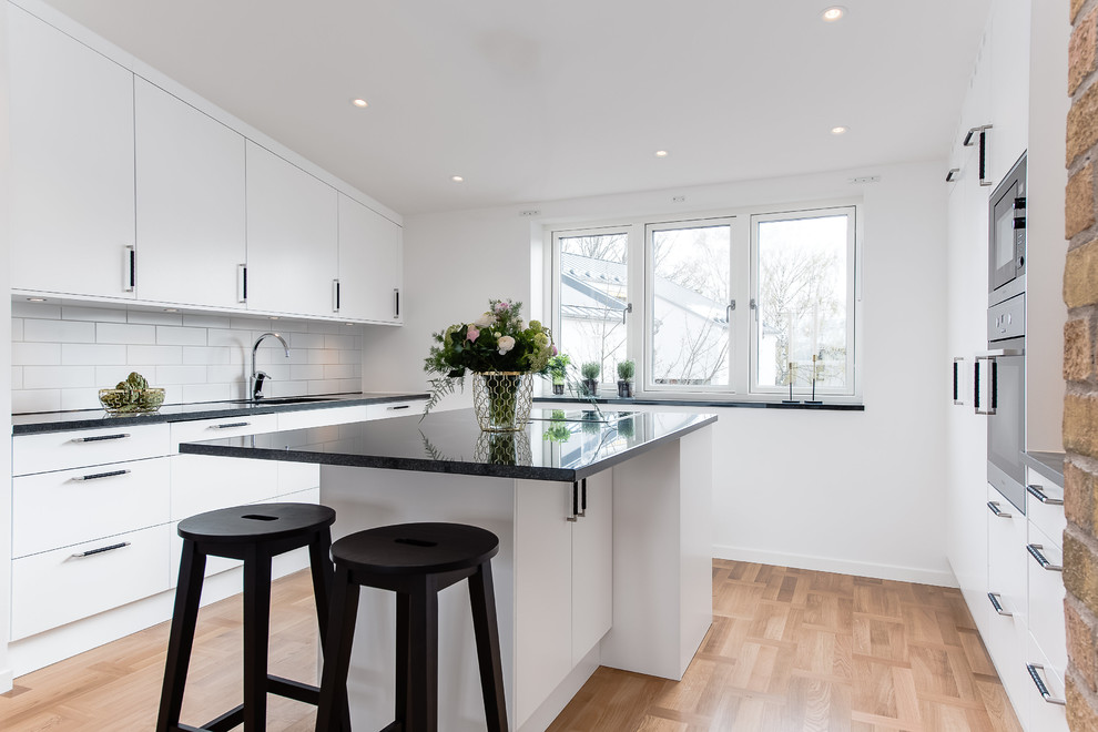 Photo of a mid-sized scandinavian single-wall kitchen in Stockholm with flat-panel cabinets, white splashback, subway tile splashback, light hardwood floors, with island and stainless steel appliances.
