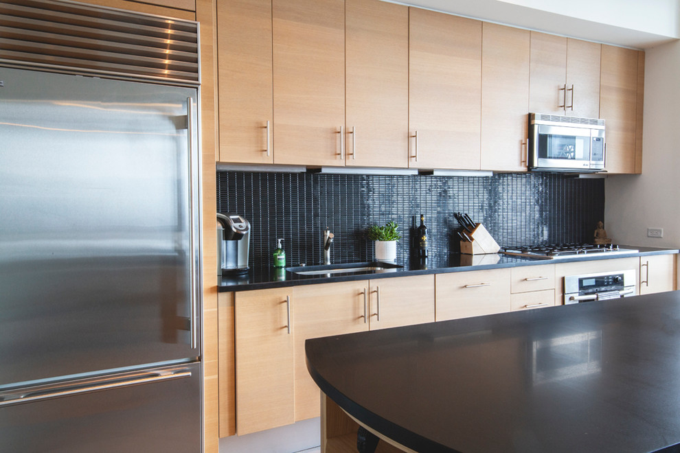 Modern single-wall open plan kitchen in New York with a drop-in sink, light wood cabinets, onyx benchtops, black splashback, mosaic tile splashback, stainless steel appliances, porcelain floors, with island, grey floor and black benchtop.