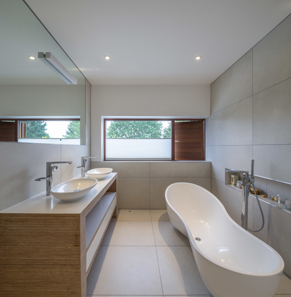 Mid-sized modern bathroom in London with a vessel sink, open cabinets, light wood cabinets, a freestanding tub, gray tile and grey walls.