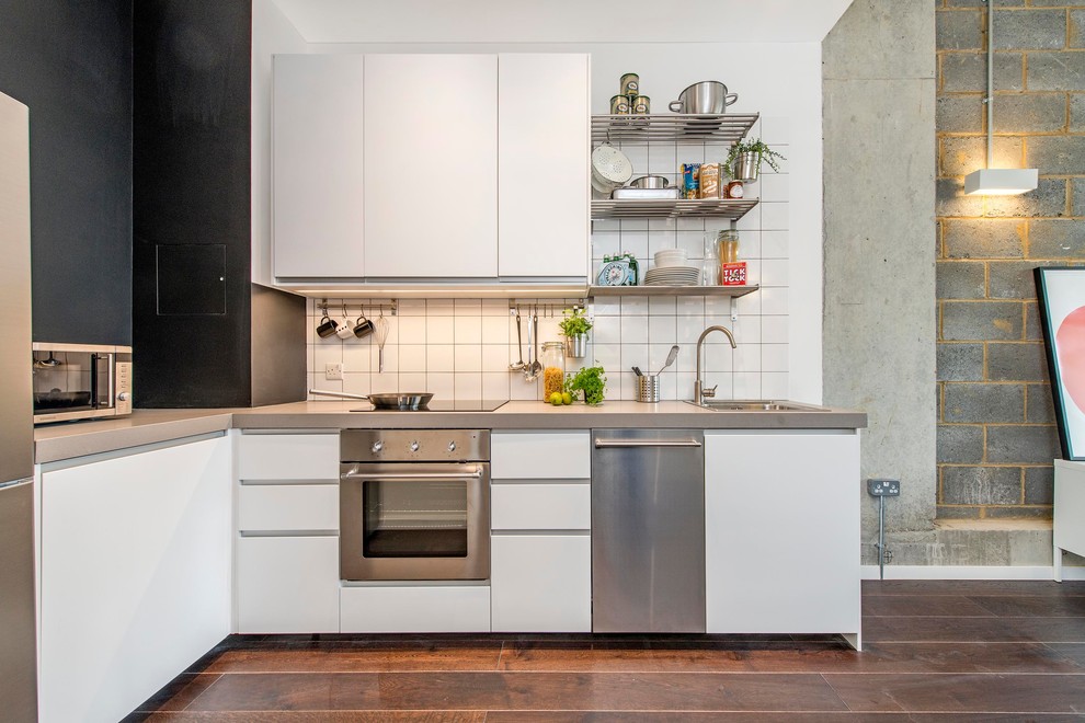 Small industrial l-shaped open plan kitchen in London with a drop-in sink, flat-panel cabinets, white cabinets, concrete benchtops, white splashback, ceramic splashback, stainless steel appliances, dark hardwood floors and no island.