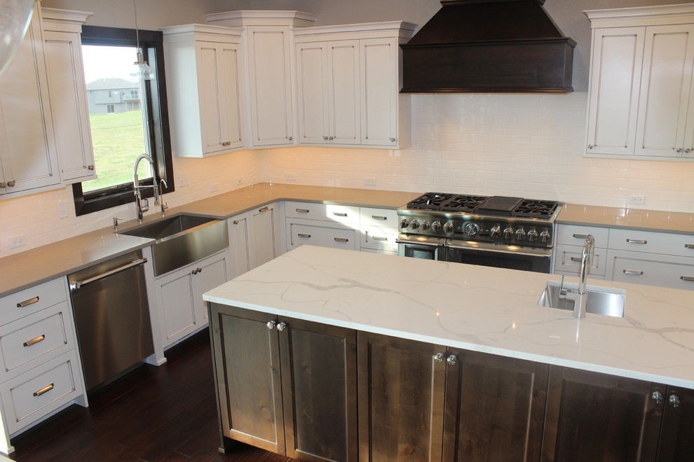 Example of a large transitional l-shaped dark wood floor enclosed kitchen design with a farmhouse sink, shaker cabinets, white cabinets, quartz countertops, white backsplash, subway tile backsplash, stainless steel appliances and an island