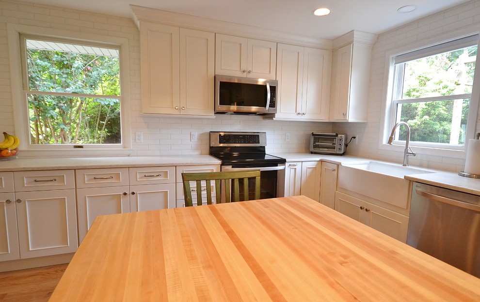 Large transitional l-shaped eat-in kitchen in Philadelphia with a farmhouse sink, shaker cabinets, white cabinets, quartz benchtops, white splashback, subway tile splashback, stainless steel appliances, light hardwood floors, with island, brown floor and white benchtop.