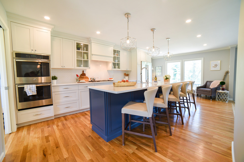 Mid-sized transitional galley open plan kitchen in Philadelphia with a farmhouse sink, shaker cabinets, white cabinets, quartz benchtops, white splashback, ceramic splashback, stainless steel appliances, medium hardwood floors, with island, brown floor and white benchtop.