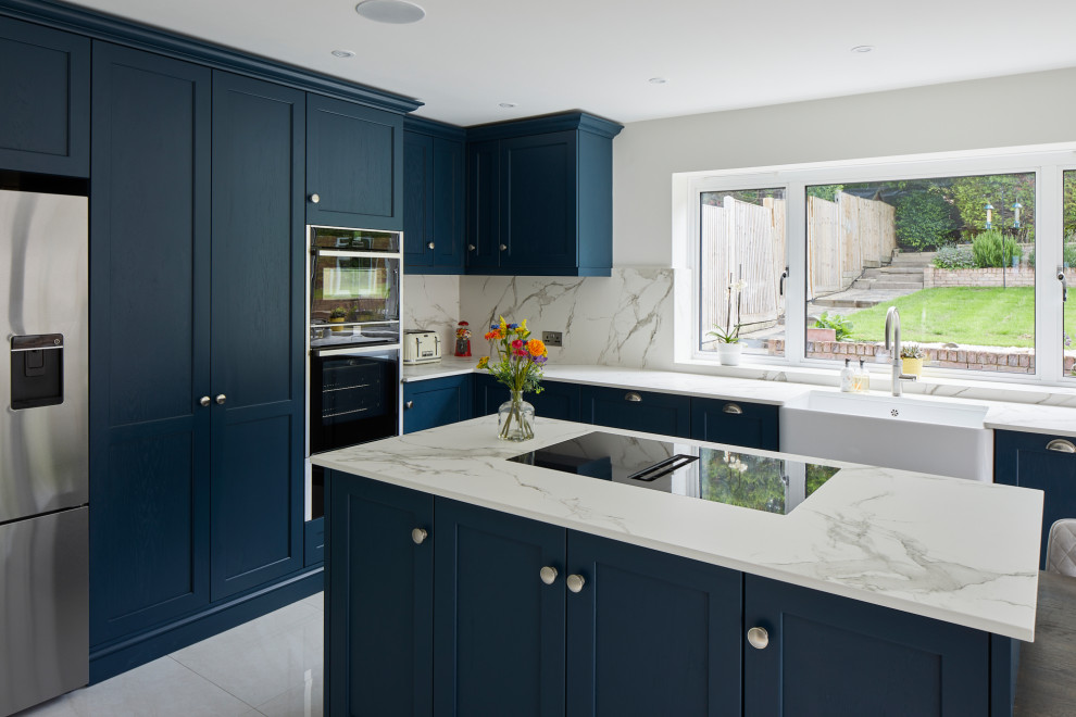 This is an example of a large contemporary l-shaped kitchen/diner in London with a belfast sink, recessed-panel cabinets, blue cabinets, engineered stone countertops, white splashback, stone slab splashback, stainless steel appliances, an island and white worktops.