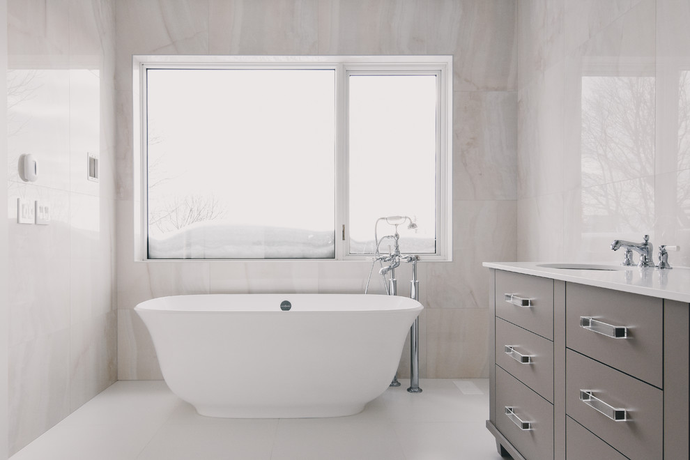 This is an example of a large contemporary master bathroom in Montreal with flat-panel cabinets, grey cabinets, a freestanding tub, white tile, porcelain tile, porcelain floors, an undermount sink and engineered quartz benchtops.