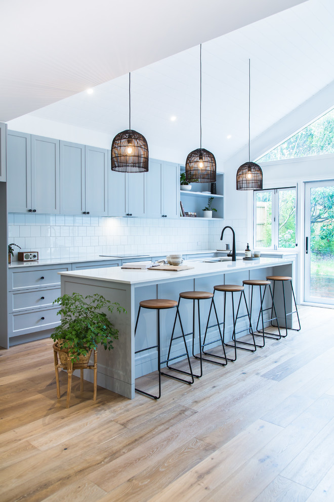 This is an example of a large modern l-shaped open plan kitchen in Melbourne with an undermount sink, shaker cabinets, grey cabinets, quartz benchtops, white splashback, ceramic splashback, black appliances, medium hardwood floors, with island, brown floor and white benchtop.