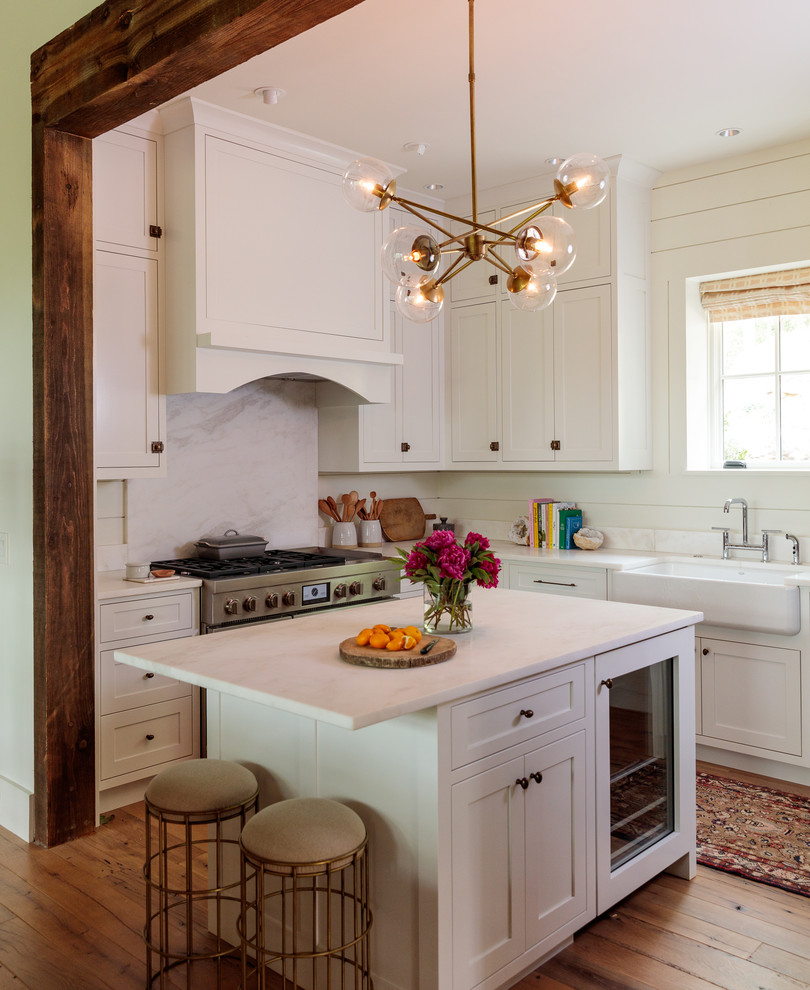 Photo of a transitional u-shaped kitchen in Other with a farmhouse sink, shaker cabinets, white cabinets, white splashback, stainless steel appliances, medium hardwood floors, with island, brown floor and white benchtop.