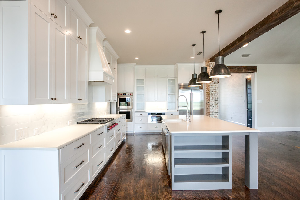 This is an example of a large country l-shaped open plan kitchen in Dallas with a farmhouse sink, shaker cabinets, white cabinets, granite benchtops, white splashback, subway tile splashback, stainless steel appliances, dark hardwood floors, with island and brown floor.