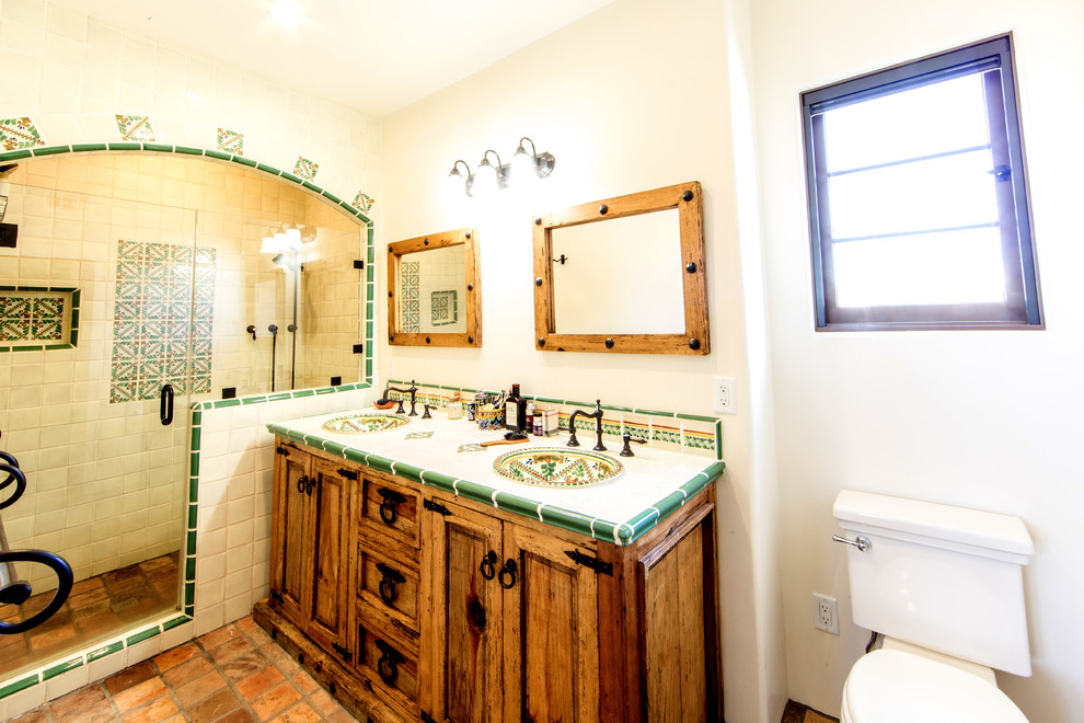 This is an example of a mid-sized mediterranean master bathroom in Los Angeles with raised-panel cabinets, brown cabinets, an alcove tub, a shower/bathtub combo, green tile, ceramic tile, white walls, a drop-in sink, tile benchtops and a hinged shower door.
