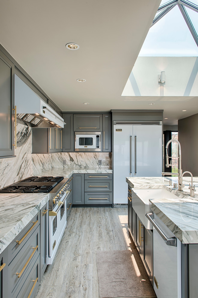 This is an example of a large transitional l-shaped eat-in kitchen in Los Angeles with a farmhouse sink, recessed-panel cabinets, grey cabinets, grey splashback, stone slab splashback, white appliances, light hardwood floors, with island, marble benchtops and beige floor.