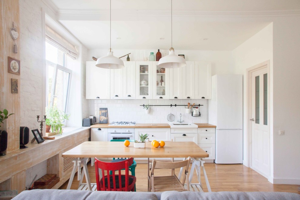 Photo of a scandinavian open plan kitchen in Other with a farmhouse sink, white cabinets, wood benchtops, white splashback, subway tile splashback, white appliances and light hardwood floors.