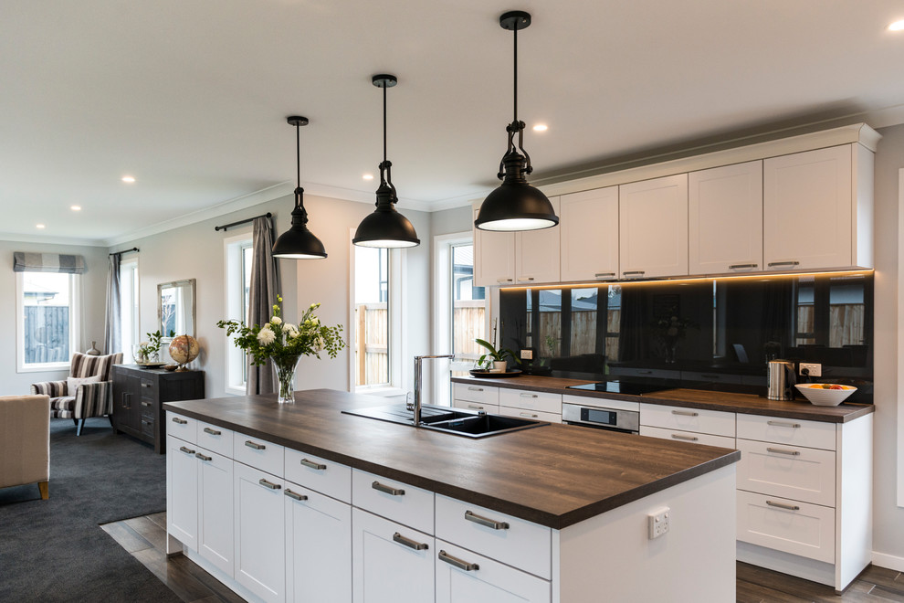 This is an example of a large transitional l-shaped kitchen pantry in Christchurch with a farmhouse sink, shaker cabinets, white cabinets, laminate benchtops, black splashback, glass sheet splashback, stainless steel appliances, vinyl floors, with island and brown floor.