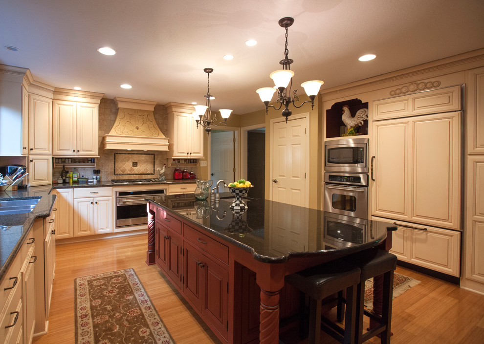 This is an example of a mid-sized traditional u-shaped eat-in kitchen in Indianapolis with an undermount sink, raised-panel cabinets, distressed cabinets, granite benchtops, beige splashback, travertine splashback, stainless steel appliances, medium hardwood floors, with island, brown floor and black benchtop.