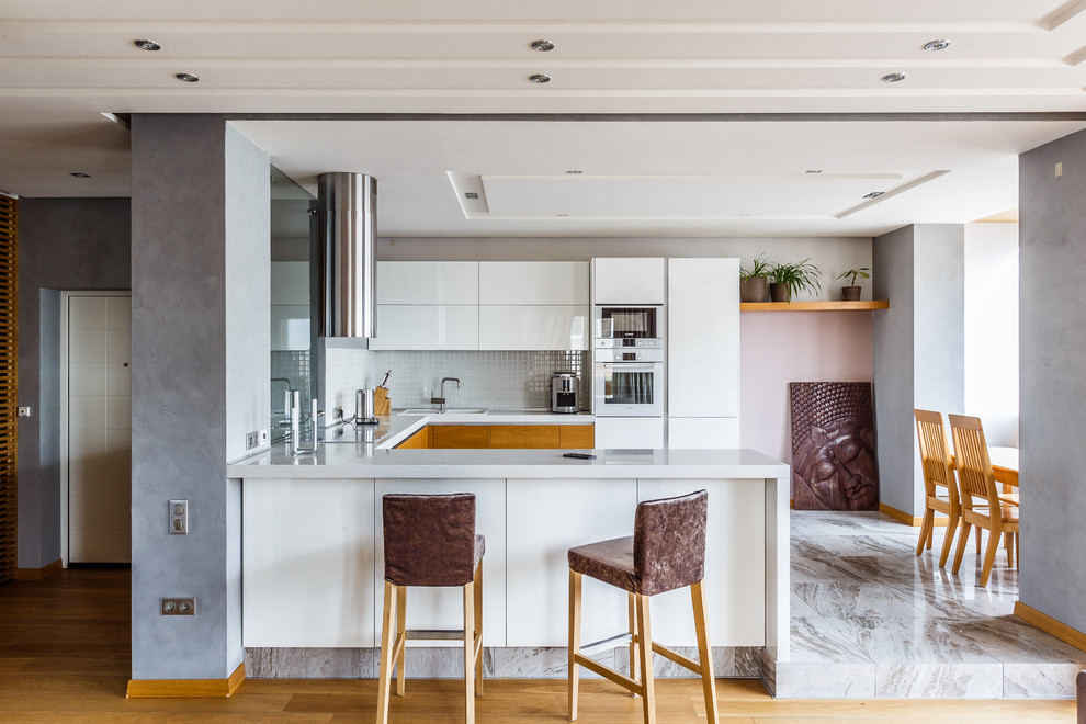 Design ideas for a mid-sized contemporary u-shaped open plan kitchen in Other with flat-panel cabinets, white cabinets, solid surface benchtops, white splashback, mosaic tile splashback, white appliances, porcelain floors, grey floor, a drop-in sink and a peninsula.